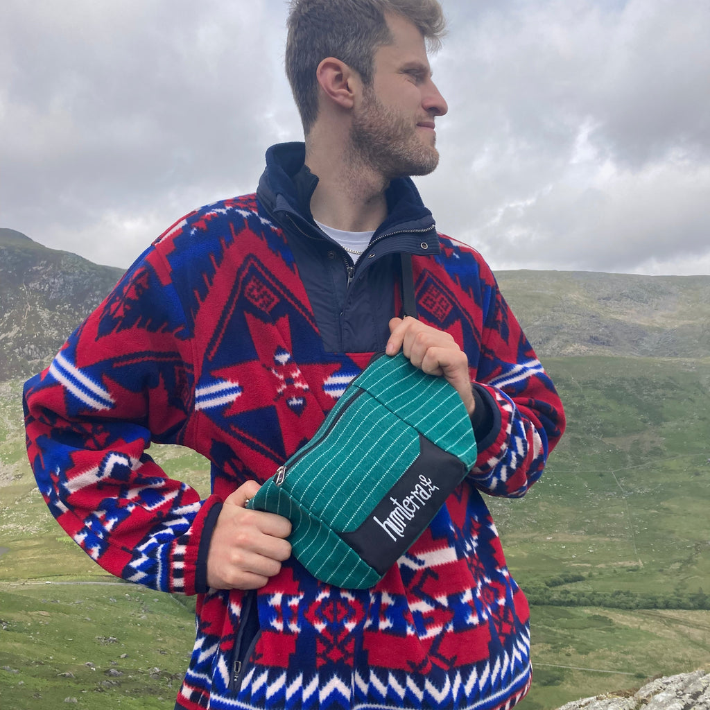 Person wearing a colourful fleece holding a green bum bag, standing against a scenic background.