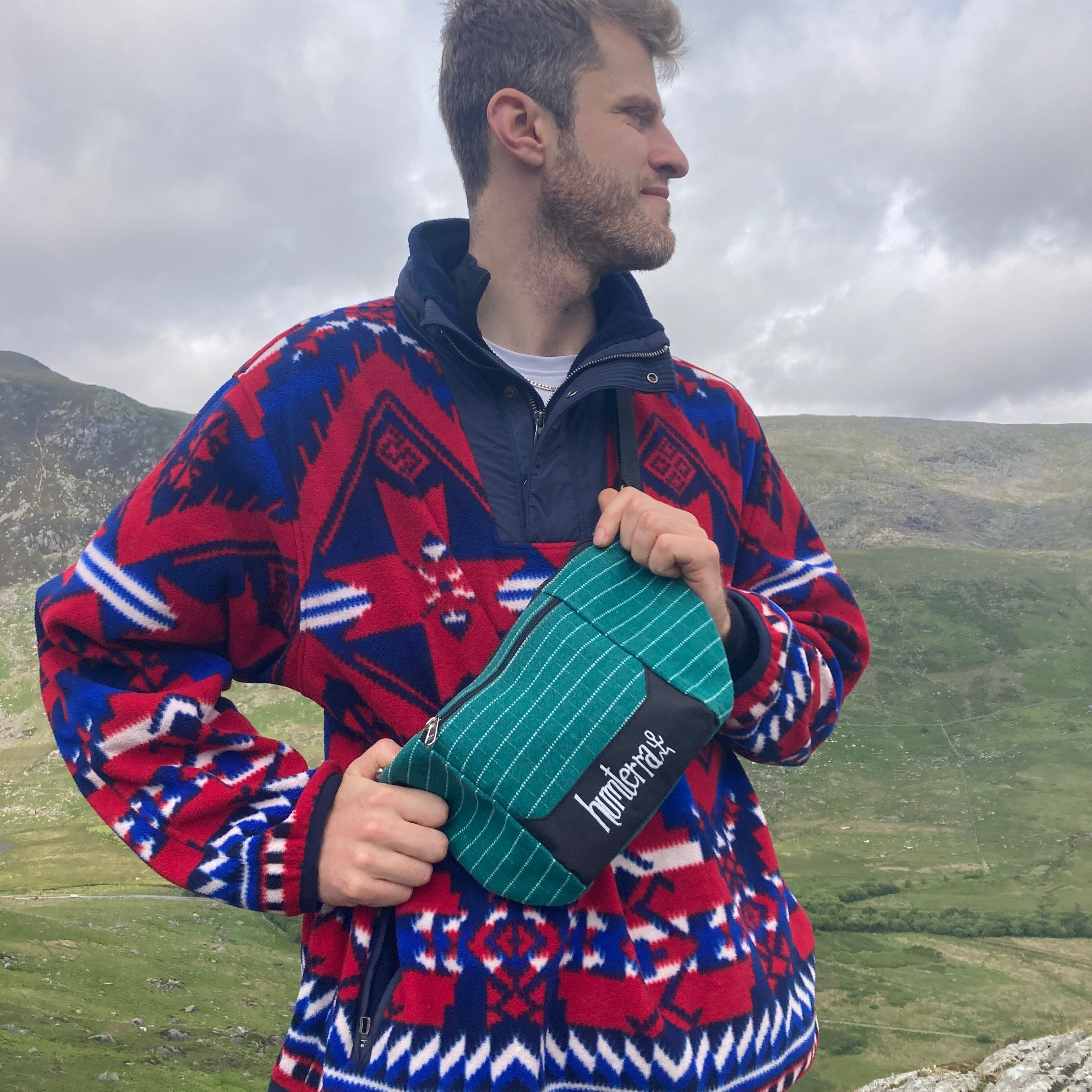 Person wearing a colourful fleece holding a green bum bag, standing against a scenic background.
