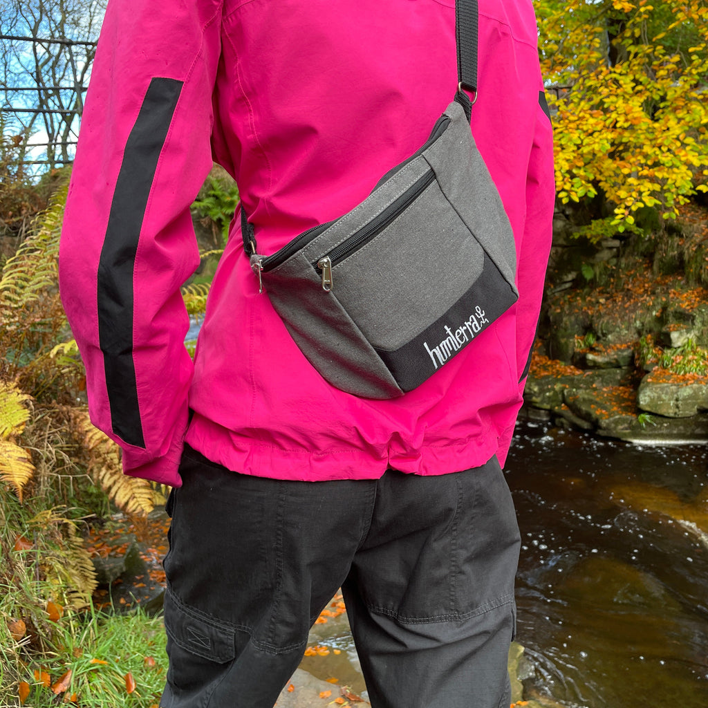 Person wearing a pink jacket with a grey fanny pack/cross body bag in a natural setting with trees and water.